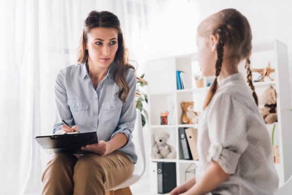 A female therapist talking to a young girl and making notes on a clipboard