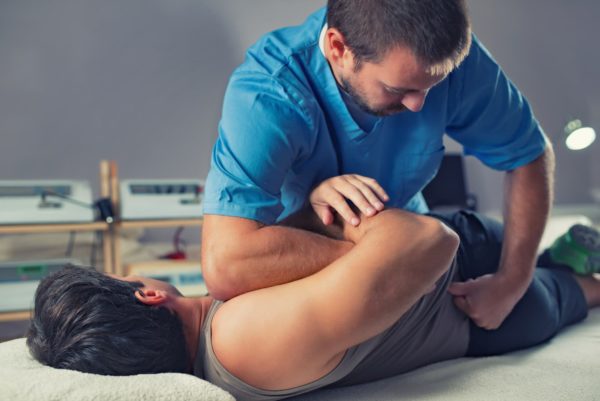A male physiotherapist is holding another man down on a table and putting pressure in the small of his back.