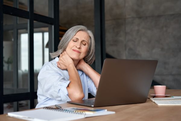 A woman sat at a desk with a laptop on it. The woman has both her hands wrapped around her neck and has her eyes closed, she looks to be in pain.