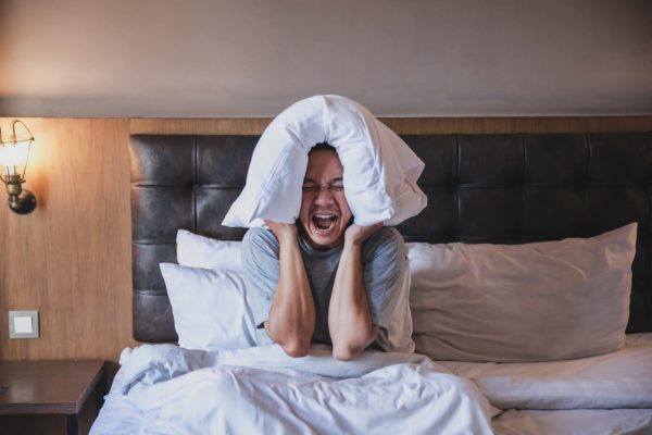 Woman sat on a bed. She is holding a pillow over the top of her head and is shouting